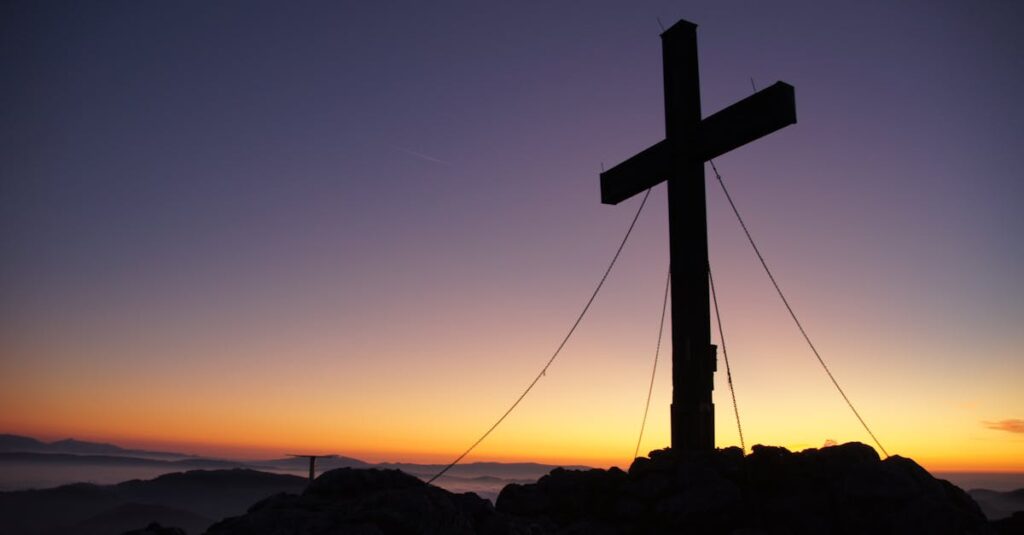 A dramatic cross stands silhouetted against a colorful sunset on a mountain peak, symbolizing faith and peace.